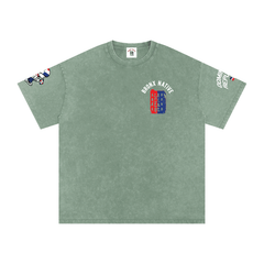 Sage green Bronx Native tee with red and blue building graphic, sleeve prints, and streetwear style design