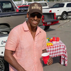 Man wearing El Bronx cap and pink shirt at outdoor tailgate with red cups and pickup trucks
