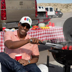 Man grilling hot dogs at outdoor BBQ, wearing cap and sunglasses, with picnic table and trucks nearby
