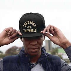 Black snapback hat with 'THE BRONX -VS- THE WORLD' in gold, worn by man in urban setting